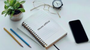Flat Lay mit Smartphone abgedreht, Lesebrille und Notizbuch, Bleistift, Armbanduhr und Pflanze – Symbol für Digital Detox im Arbeitsalltag.